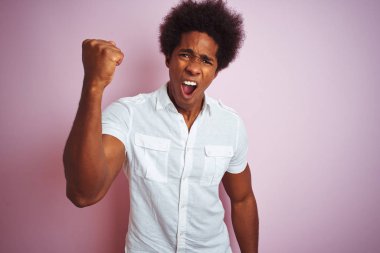 Afro saçlı, beyaz tişörtlü genç bir adam izole edilmiş pembe arka plan üzerinde öfkeli ve kızgın duruyor öfkeyle bağırırken yumruğunu kaldırıyor. Öfke ve agresif kavram.