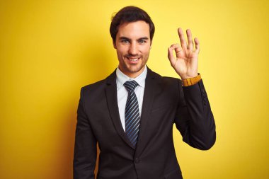 Takım elbiseli ve kravatlı genç yakışıklı işadamı izole edilmiş sarı arka planda duruyor. Gülümsüyor ve el ve parmaklarıyla 