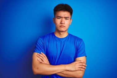 İzole edilmiş mavi arka plan üzerinde duran Asyalı genç adam şüpheci ve gergin, kollarını kavuşturmuş yüz ifadesini onaylamıyor. Negatif kişi.
