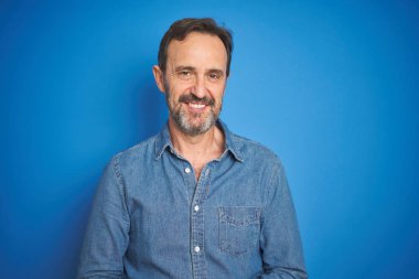 Yakışıklı, orta yaşlı, gri saçlı, soyutlanmış mavi arka planı olan, elleri çapraz parmaklı, gevşemiş ve neşeli gülümseyen bir adam. Başarı ve iyimser