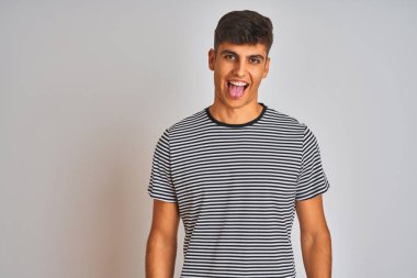 Donanma çizgili tişört giyen genç Hintli adam izole edilmiş beyaz arka planda dikiliyor ve komik bir ifadeyle dilini dışarı çıkarıyor. Duygu kavramı.