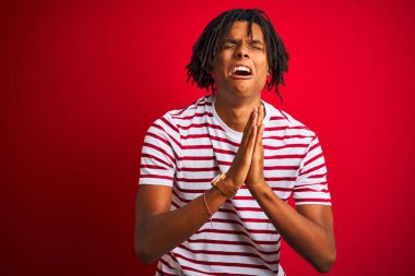Rastalı tişörtlü genç afro bir adam izole edilmiş kırmızı arka plan üzerinde duruyor umut dolu bir ifadeyle birlikte çok duygusal ve endişeli bir şekilde yalvarıyor ve dua ediyor. Af diliyor. Din kavramı.