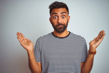 Siyah çizgili tişört giyen genç Hintli adam izole edilmiş beyaz arka plan üzerinde duruyor. Kolları ve elleri havada, kafası karışmış bir ifade. Şüphe kavramı.