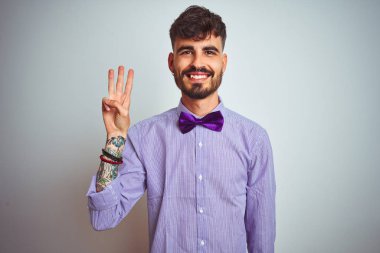 Mor gömlekli ve papyonlu genç adam izole edilmiş beyaz arka planda kendini beğenmiş ve mutlu gülümserken üç numaralı parmaklarıyla işaret ediyor..
