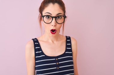 Kızıl saçlı, çizgili tişörtlü bir kadın izole edilmiş pembe arka planda dikiliyor. Şaşırmış bir yüz ifadesinden korkuyor ve heyecanlanıyor.