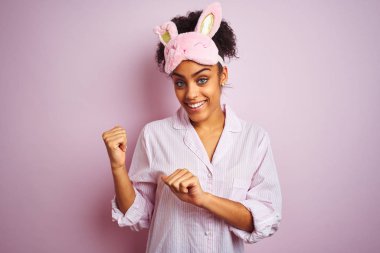 Genç Afro-Amerikalı kadın pijama giyiyor ve maske takıyor. Pembe arka planda. Arka tarafı işaret ediyor.