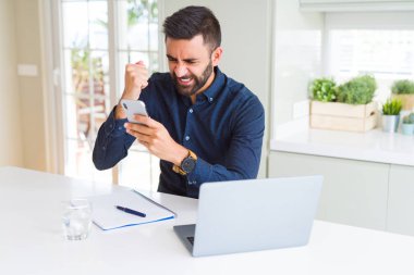 Ofiste akıllı telefon ve dizüstü bilgisayar kullanan yakışıklı İspanyol iş adamı sinirlenmiş ve sinirlenmiş bağırıyor, çıldırmış ve el kaldırarak bağırıyor. Öfke kavramı.