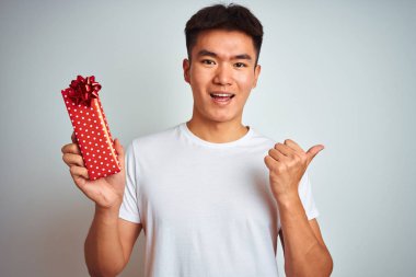 Sevgililer günü kartı tutan Asyalı genç adam izole edilmiş beyaz arka planda duruyor ve gülümseyen bir gülümsemeyle baş parmağıyla onu işaret ediyor.
