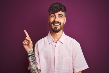 Yalıtılmış mor arka plan üzerinde duran pembe tişörtlü genç adam yüzünde kocaman bir gülümseme, eli ve parmağı kameraya bakıyor..