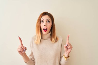 Kızıl saçlı kadın zarif balıkçı yaka kazak giyiyor. İzole edilmiş beyaz arka plan üzerinde duruyor. Şaşırmış ve şaşırmış bir şekilde yukarı bakıyor ve parmaklarıyla işaret ediyor..