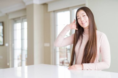 Güzel Asyalı kadın, beyaz masada rahat bir süveter giyip gülümsüyor telefonla el ve parmaklarıyla jest yapıyor telefonda konuşuyor gibi. İletişim kavramları.