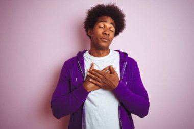 Mor eşofman altı giyen genç Afro-Amerikalı adam izole edilmiş pembe arka planda duruyor elleri göğsünde, gözleri kapalı ve yüzünde minnet dolu bir jest ile gülümsüyor. Sağlık kavramı.