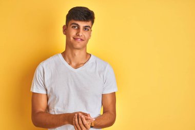 İzole edilmiş sarı arka plan üzerinde duran beyaz tişörtlü genç Hintli adam ellerini kavuşturup gülümseyerek gevşemiş ve neşeli gülümsüyor. Başarı ve iyimser