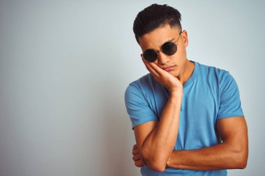 İzole edilmiş beyaz arka planda mavi tişört ve güneş gözlüğü takan genç Brezilyalı adam, çapraz kollu depresyon sorunlarından sıkılmış ve yorgun görünüyor..