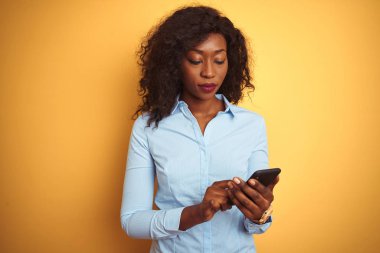 Genç Afro-Amerikalı kadın akıllı telefon kullanıyor. İzole edilmiş sarı arka planda duruyor. Akıllı bir yüz ifadesi var.