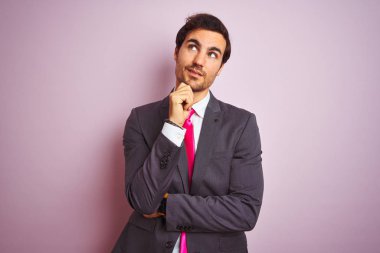Takım elbiseli ve kravatlı genç yakışıklı bir işadamı pembe arka planda, elleri çenesinde soru sorarken, düşünceli bir ifade takınıyor. Düşünceli bir yüzle gülümsüyor. Şüphe kavramı.