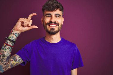 İzole edilmiş mor arka plan üzerinde duran tişörtlü genç adam gülümsüyor ve el hareketleriyle küçük boy işareti yapıyor ve kameraya bakıyor. Ölçü kavramı.
