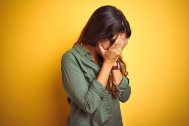 Sarı arka plan üzerinde yeşil gömlek ve gözlük takan genç ve güzel bir kadın. Ağlarken suratını ellerle kapatan üzgün bir yüz ifadesi. Depresyon kavramı.