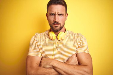 Kulaklık takan genç yakışıklı bir adam sarı izole edilmiş arka planda müzik dinliyor şüpheci ve gergin, yüzünde çapraz kollarla onaylamayan bir ifade var. Negatif kişi.