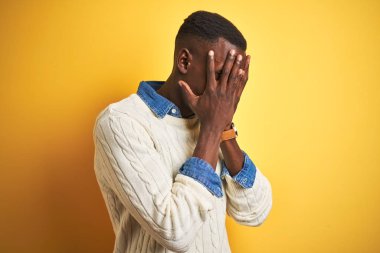 Kotlu gömlek ve beyaz süveter giyen Afro-Amerikan bir adam izole edilmiş sarı arka plan ve ağlarken suratını ellerle kapatan üzgün bir yüz ifadesi. Depresyon kavramı.