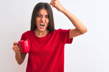 Genç ve güzel bir kadın izole edilmiş beyaz arka planda kırmızı kahve içerken sinirlenmiş ve sinirlenmiş bir şekilde bağırıyor, çıldırmış ve el kaldırarak bağırıyor.