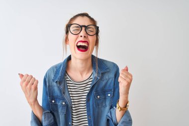 Çizgili tişört ve gözlük takan kızıl saçlı kadın izole edilmiş beyaz arka planda çok mutlu ve heyecanlı bir şekilde kollarını kaldırıp, gülümseyerek ve başarı için çığlık atarak zafer hareketi yapıyor. Kutlama konsepti.