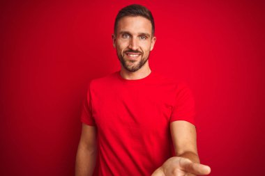 Kırmızı izole edilmiş arka plan üzerine gündelik tişört giyen genç yakışıklı adam gülümseyerek avuçlarını açıp yardım ve kabullenme teklif ediyor..