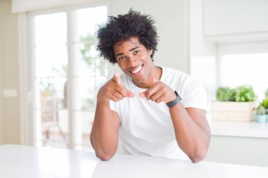 Genç Afro-Amerikalı adam, günlük beyaz tişörtler giyip evde oturmuş, seni ve kamerayı parmaklarıyla göstererek, neşeli ve pozitif gülümsüyor.