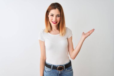 Izole beyaz arka plan üzerinde duran rahat t-shirt giyen güzel kızıl saçlı kadın neşeli sunan gülümseyerek ve kameraya bakarak avuç içi ile işaret.