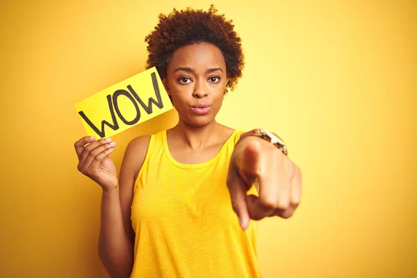 Young african american woman showing wow banner board over yellow isolated background pointing with finger to the camera and to you, hand sign, positive and confident gesture from the front