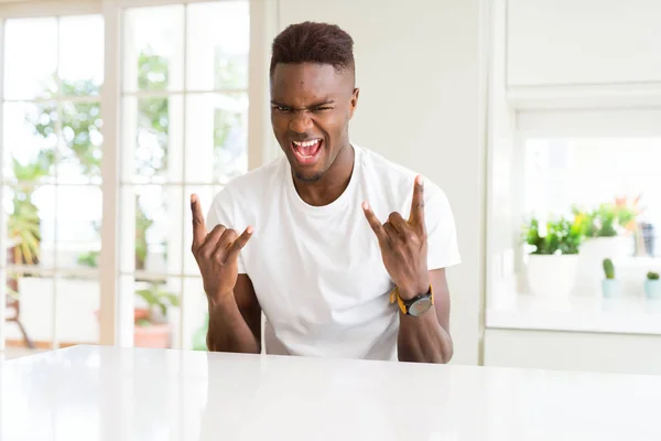 一个英俊的非洲裔美国男人在家里的白桌子上大声喊着 用疯狂的表情举手表决摇滚符号 音乐明星 沉重的概念 — 图库照片