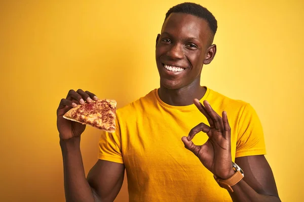 African american man eating slice of pizza standing over isolated yellow background doing ok sign with fingers, excellent symbol