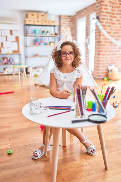 Beautiful toddler wearing glasses sitting drawing using paper and pencils at kindergarten