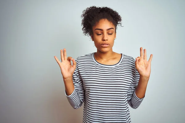 African american woman wearing navy striped t-shirt standing over isolated white background relaxed and smiling with eyes closed doing meditation gesture with fingers. Yoga concept.