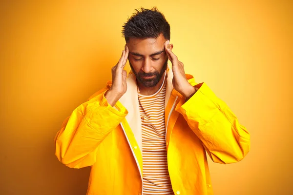 Young indian man wearing raincoat standing over isolated yellow background with hand on head for pain in head because stress. Suffering migraine.