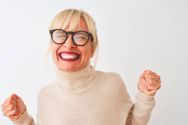 Middle age woman wearing turtleneck sweater and glasses over isolated white background very happy and excited doing winner gesture with arms raised, smiling and screaming for success. Celebration concept.