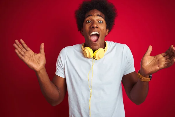 年轻的非洲裔美国人在孤立的红色背景下 用耳机听着音乐 欢呼着疯狂的成功 欢呼着张开双臂 睁开眼睛 兴奋地欢呼着 赢的概念 — 图库照片