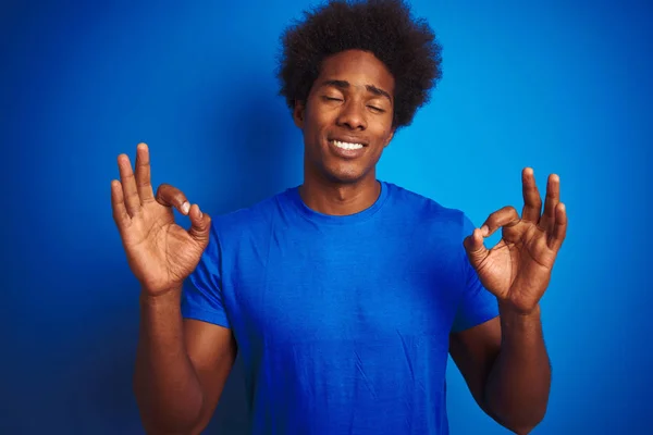 African american man with afro hair wearing t-shirt standing over isolated blue background relax and smiling with eyes closed doing meditation gesture with fingers. Yoga concept.