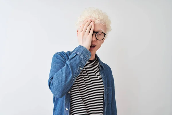 Young albino blond man wearing denim shirt and glasses over isolated white background covering one eye with hand, confident smile on face and surprise emotion.