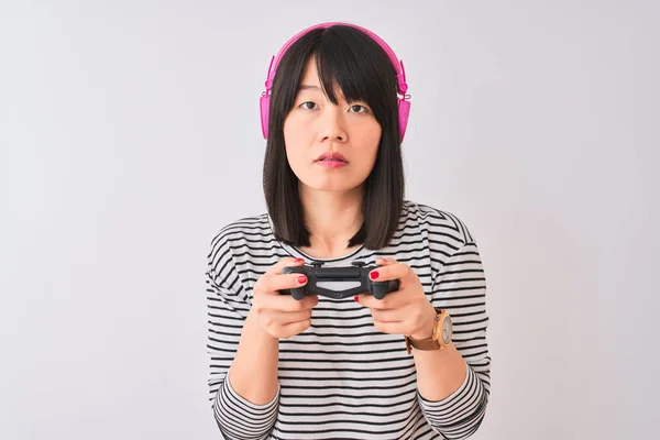 Chinese gamer woman playing video game using headphones over isolated white background with a confident expression on smart face thinking serious