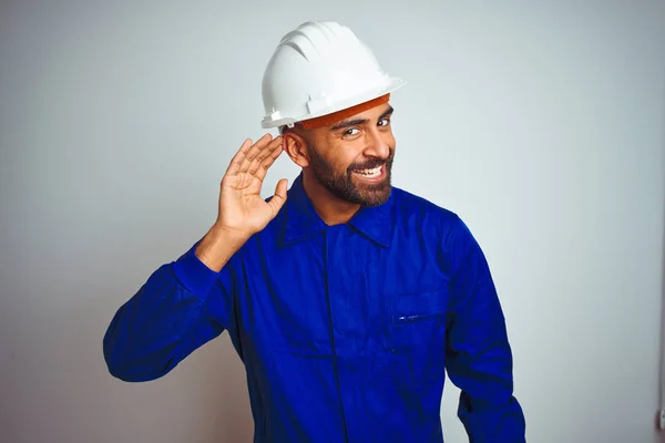 Handsome indian worker man wearing uniform and helmet over isolated white background smiling with hand over ear listening an hearing to rumor or gossip. Deafness concept.