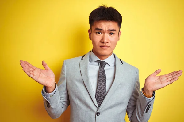 Asian chinese businessman wearing suit and tie standing over isolated yellow background clueless and confused expression with arms and hands raised. Doubt concept.