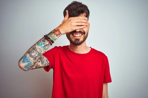 Young man with tattoo wearing red t-shirt standing over isolated white background smiling and laughing with hand on face covering eyes for surprise. Blind concept.