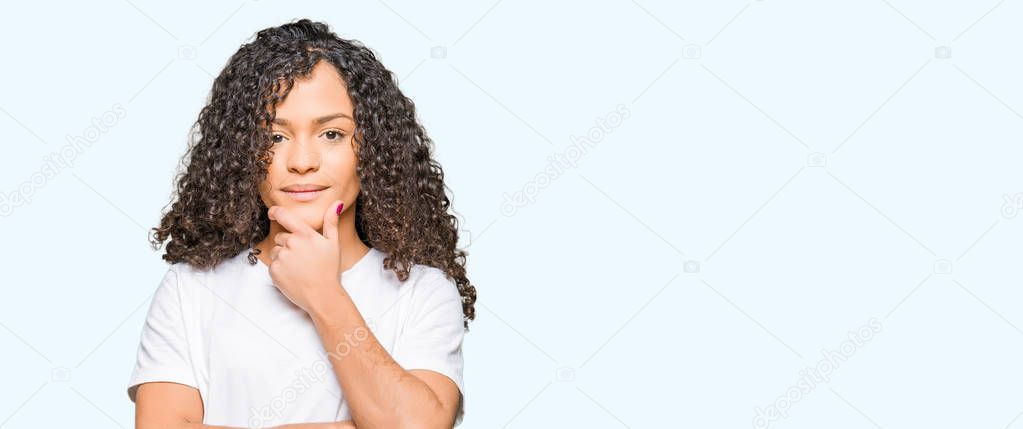 Young beautiful woman with curly hair wearing white t-shirt looking confident at the camera with smile with crossed arms and hand raised on chin. Thinking positive.