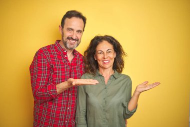 İzole sarı arka plan üzerinde güzel orta yaş çift gülümseyen neşeli sunumu ve kamera bakarak el avuç ile işaret.