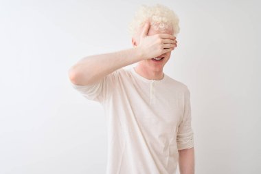 Sıradan tişörtler giyen genç albino bir adam izole edilmiş beyaz arka planda dikiliyor, gülümsüyor ve yüzünde şaşkınlık için gözleri kapayarak gülüyor. Kör kavram.