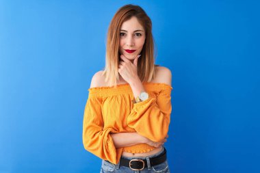 Güzel kızıl saçlı kadın turuncu günlük tişört giyiyor mavi arka planda dikiliyor kameraya bakıyor kollarını kavuşturup gülümseyerek ve çenesini kaldırarak bakıyor. Olumlu düşünüyorum..