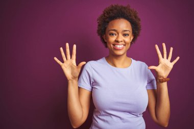 Afro saçlı, güzel, Afro saçlı genç bir Amerikalı kadın. Mor arka planda on numarayı işaret ediyor. Kendinden emin ve mutlu bir şekilde gülümsüyor..