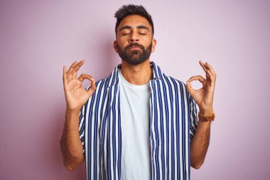 Genç ve yakışıklı Hintli adam yaz çizgili gömleğini pembe arka planda giyiyor rahatla ve gözleri kapalı gülümse parmaklarıyla meditasyon hareketi yapıyor. Yoga konsepti.