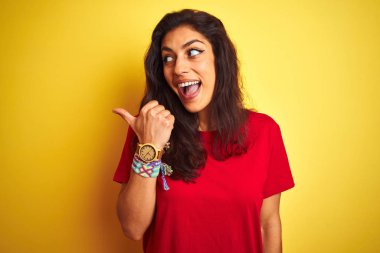 Kırmızı tişörtlü genç bir kadın izole edilmiş sarı arka planda dikiliyor ve baş parmağıyla gülümsüyor.